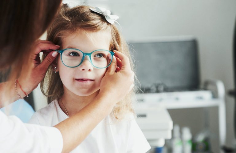 Ein Kind mit blauer Brille dem von einer Person beim Anpassen der Brille geholfen wird.