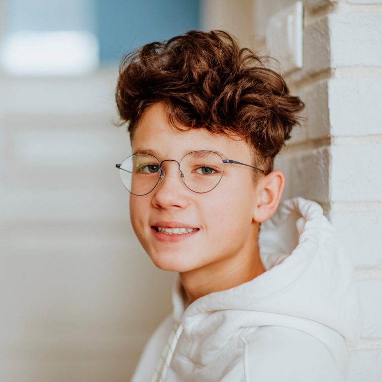 Junger Mann mit lockigem Haar und Brille, lächelt in einer hellen Umgebung.