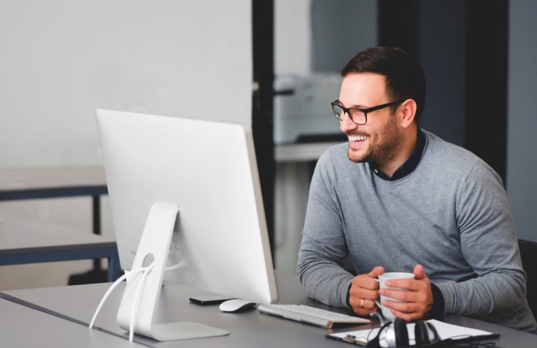 Lächelnder Mann mit Officebrille sitzt an einem Schreibtisch und schaut auf einen Computerbildschirm.