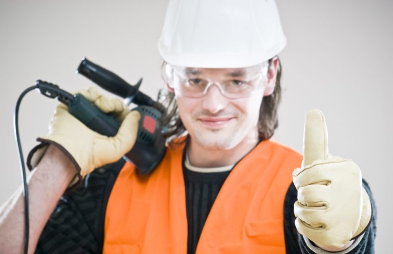 Mann in Sicherheitskleidung mit Schutzbrille, hält ein Werkzeug und zeigt Daumen hoch.